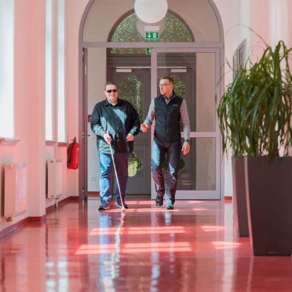 Bildbeschreibung: Zwei HWG Mitarbeitende laufen im Flur. Ein Mann hat einen Langstock und läuft links.