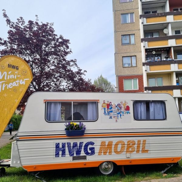 Bildbeschreibung: Wohnwagenanhänger mit Aufschrift HWG Mobil steht vor einem Plattenbaublock.